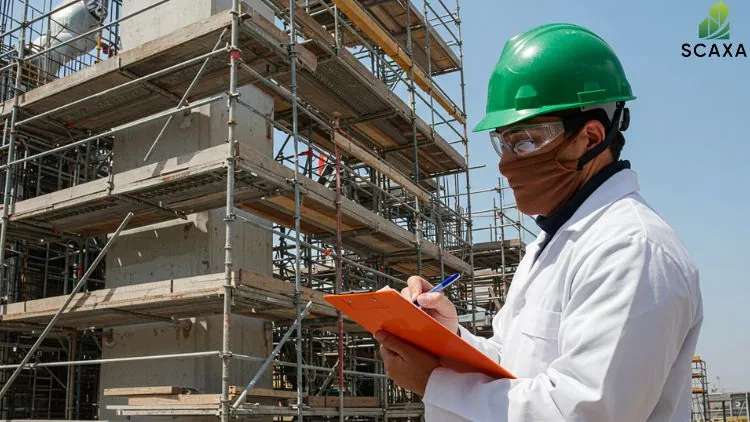 Construction worker in a green hard hat and safety goggles, holding a clipboard, inspecting scaffolding structures at a construction site, with SCAXA branding visible.